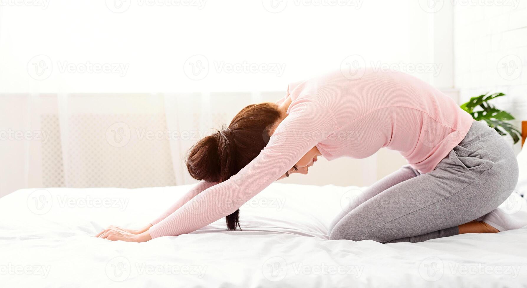 A woman in a bedroom is stretching her body on a bed, extending her arms and legs in different directions. The room is neatly arranged with a bed, pillows, and a bedside table, side view photo