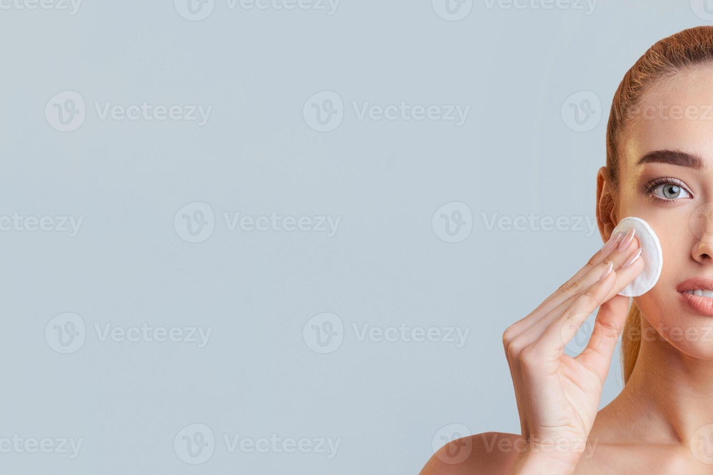 This image shows a woman with fair skin applying makeup remover to her face with a cotton pad. She is looking at the camera and has her hair pulled back in a ponytail, copy space photo