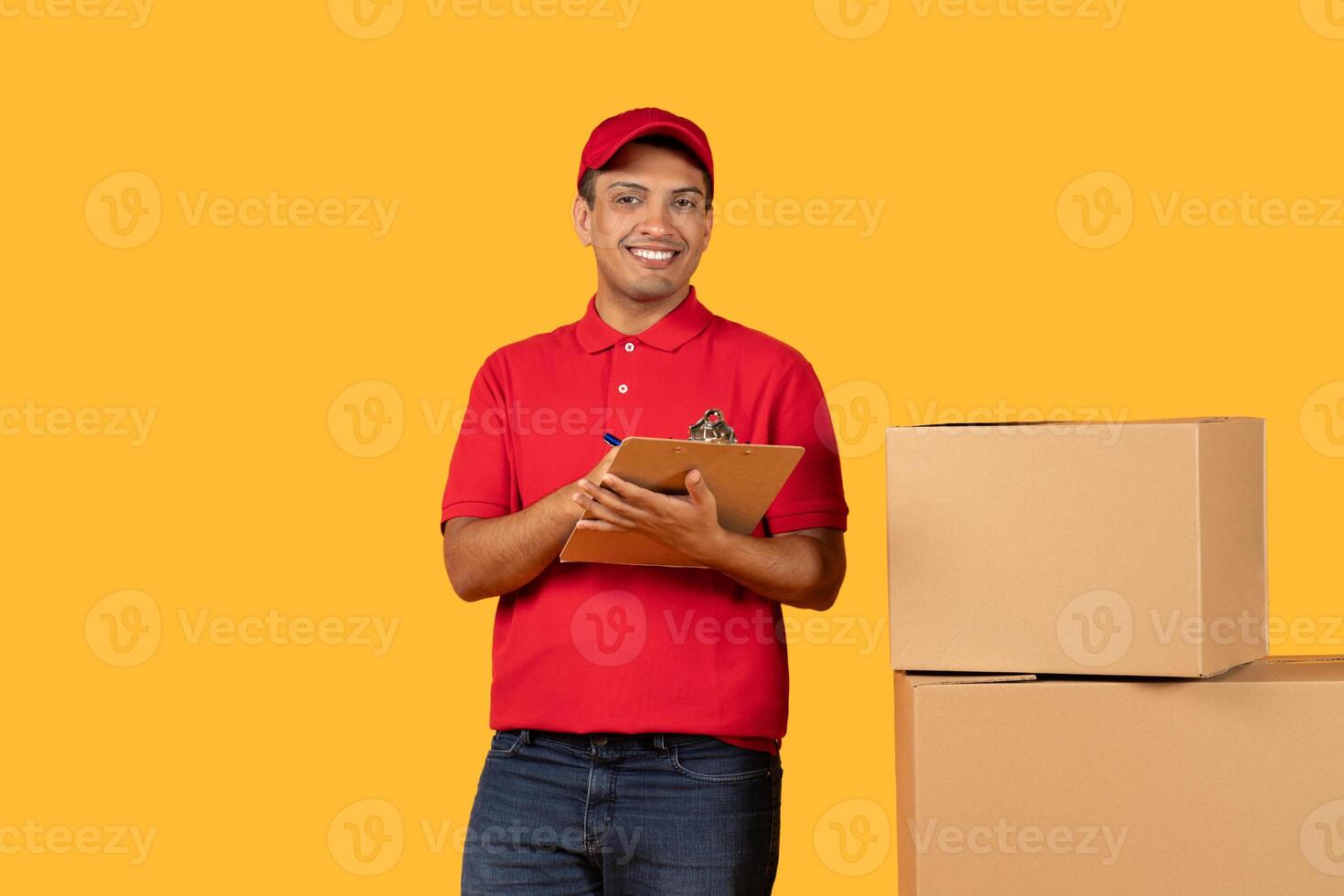 A cheerful delivery guy in a red uniform holds a clipboard while standing beside cardboard boxes. The bright yellow background highlights his friendly demeanor as he prepares for a package delivery. photo