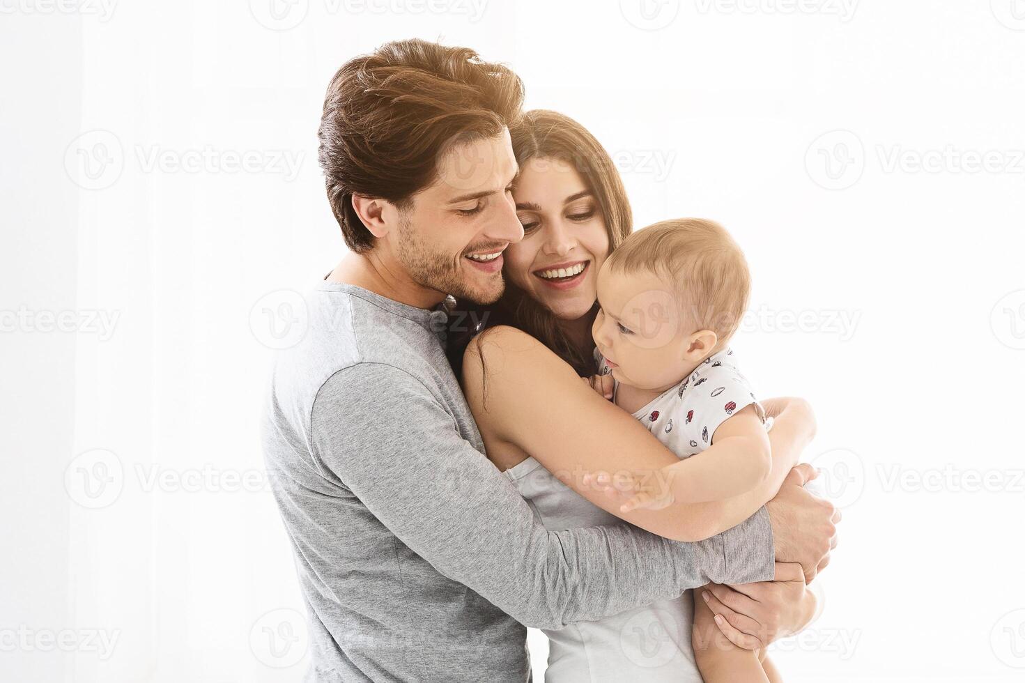 This image shows a happy family of three, a man, a woman, and their baby, standing close together in a white interior setting. The man and woman are smiling at the baby photo