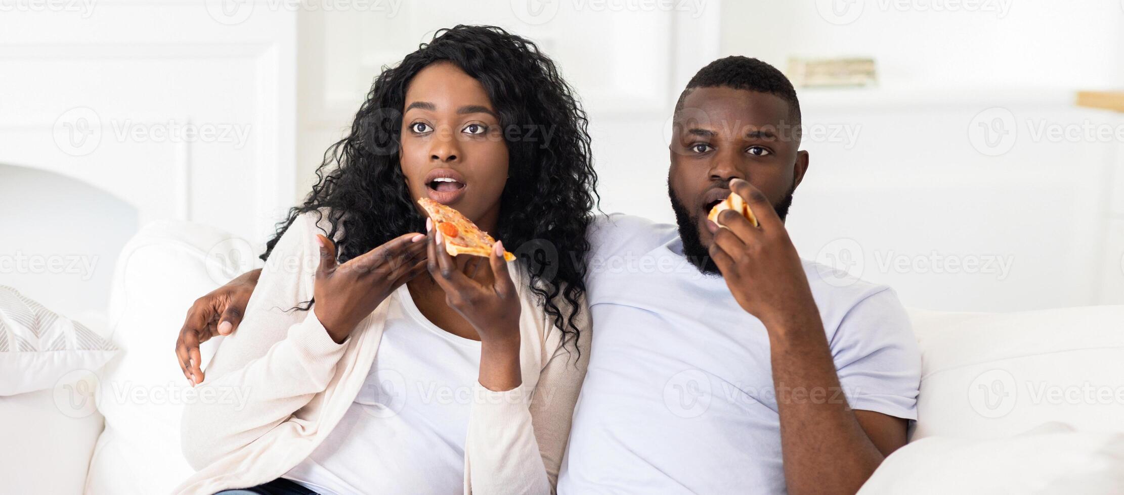 A couple enjoys a slice of pizza while relaxing on a white couch. They look to be enjoying their meal. photo