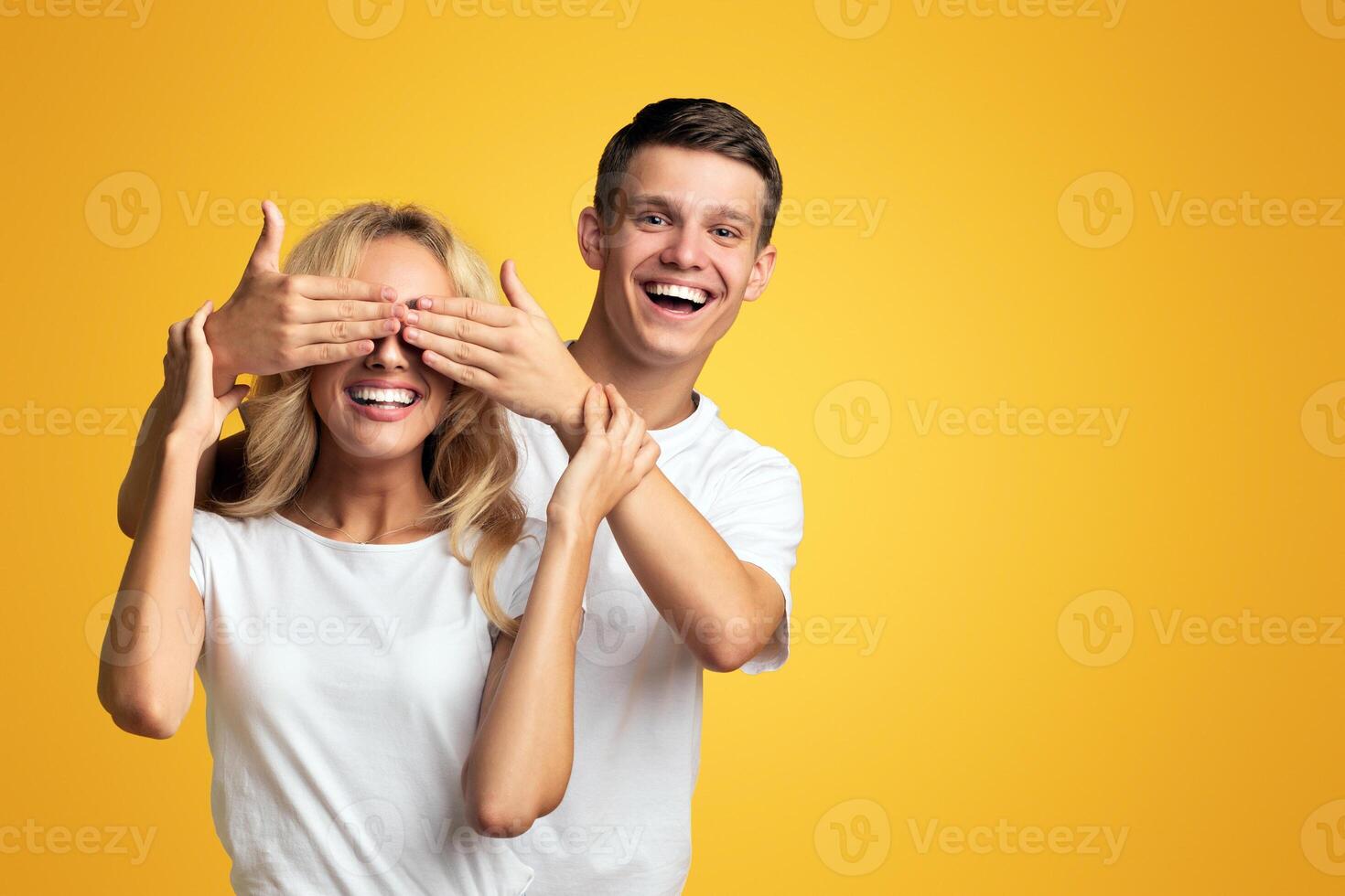 Guess who. Playful young guy standing behind girlfriend and closing her eyes, yellow studio background with copy space photo