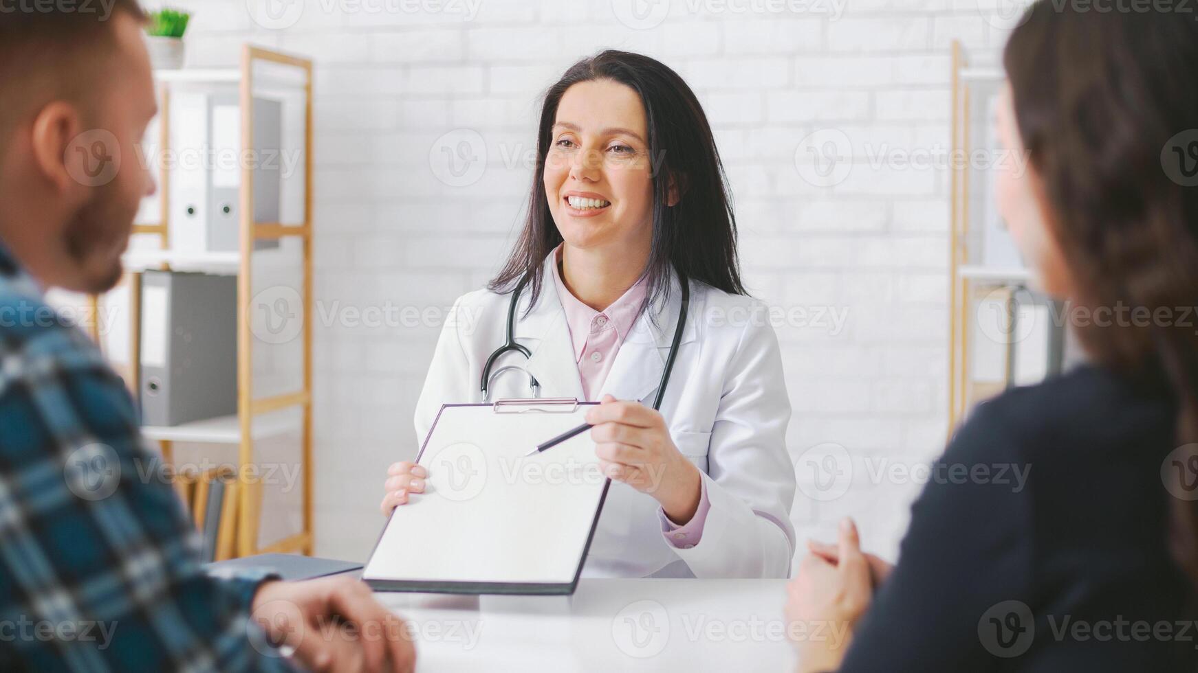 Woman doctor wearing a white coat is sitting at a table and discussing medical information with a couple. She is holding a clipboard and pointing at it with a pen. photo