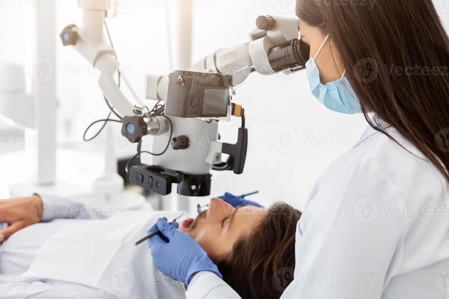 Female professional dentist doctor making different treatment in modern cabinet, using microscope, side view photo