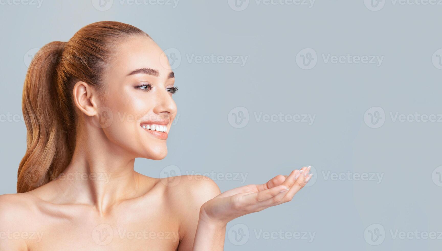 A woman with brown hair tied up in a ponytail is smiling and holding her hand out in front of her. She is standing in front of a light blue background. Her skin is clear and smooth, copy space photo