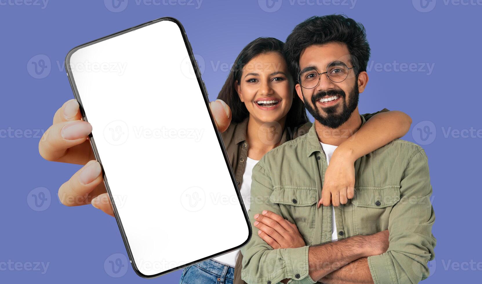 A young Indian couple is standing in front of a blue background, holding a smartphone with a blank screen. The woman is smiling and has her arm around the man's shoulders. photo