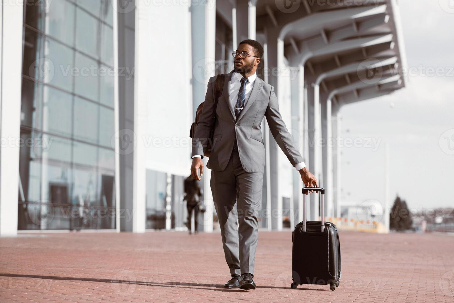 Black businessman walking with rolling suitcase outside a modern building, ready for a business trip or travel. photo