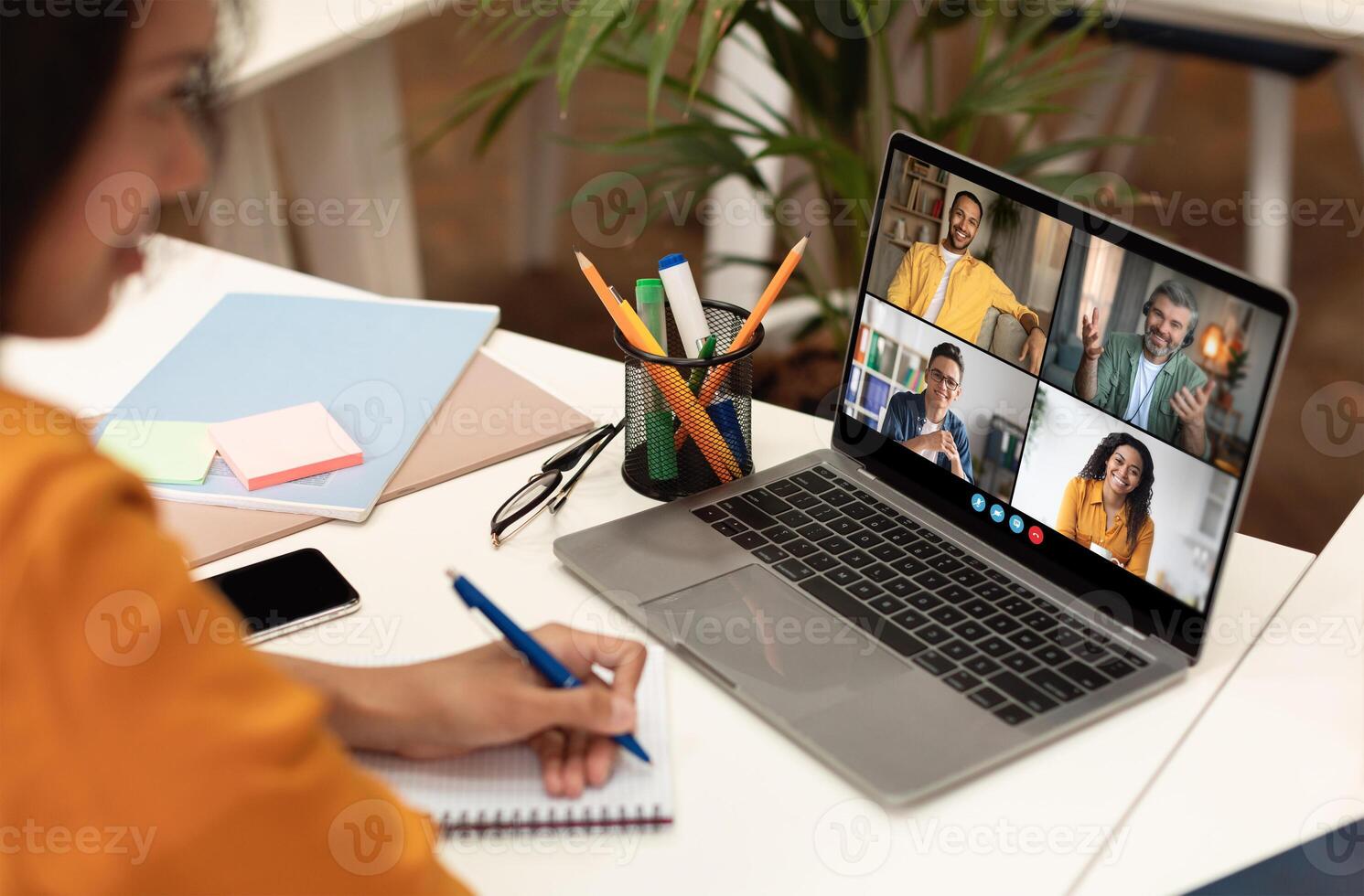 A woman is sitting at a desk, taking notes in a notebook while participating in a conference on a laptop. The laptop screen displays four participants in a call photo