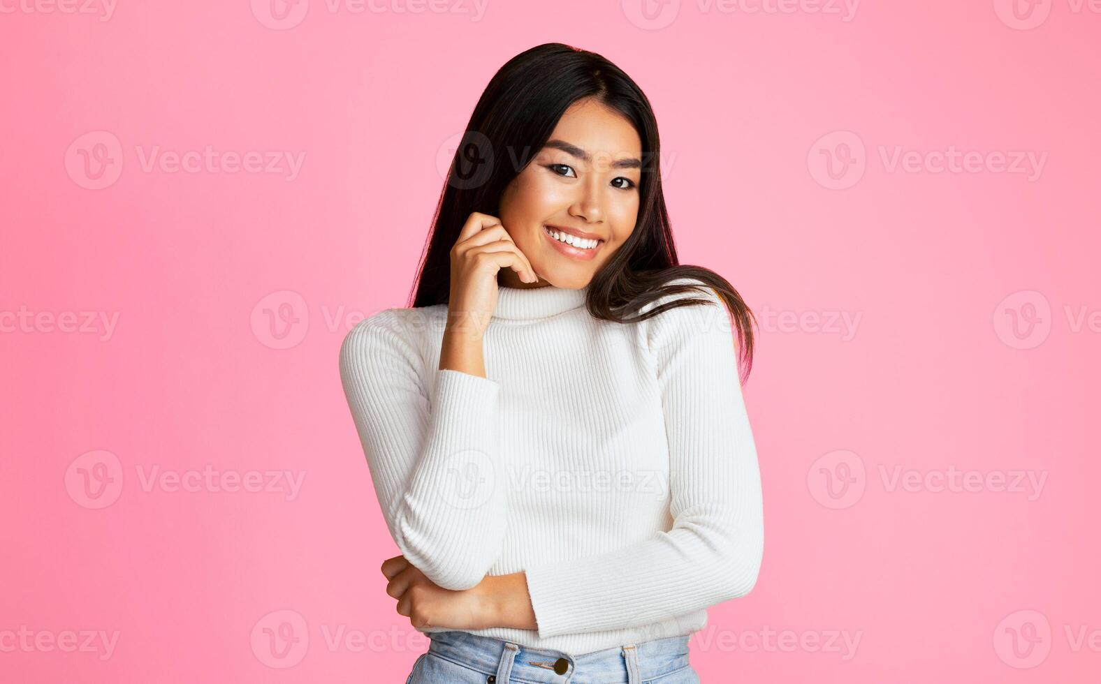 Asian young woman with long black hair is smiling and looking at the camera. She is wearing a white sweater and has her hand on her chin. The background is a solid pink color. photo