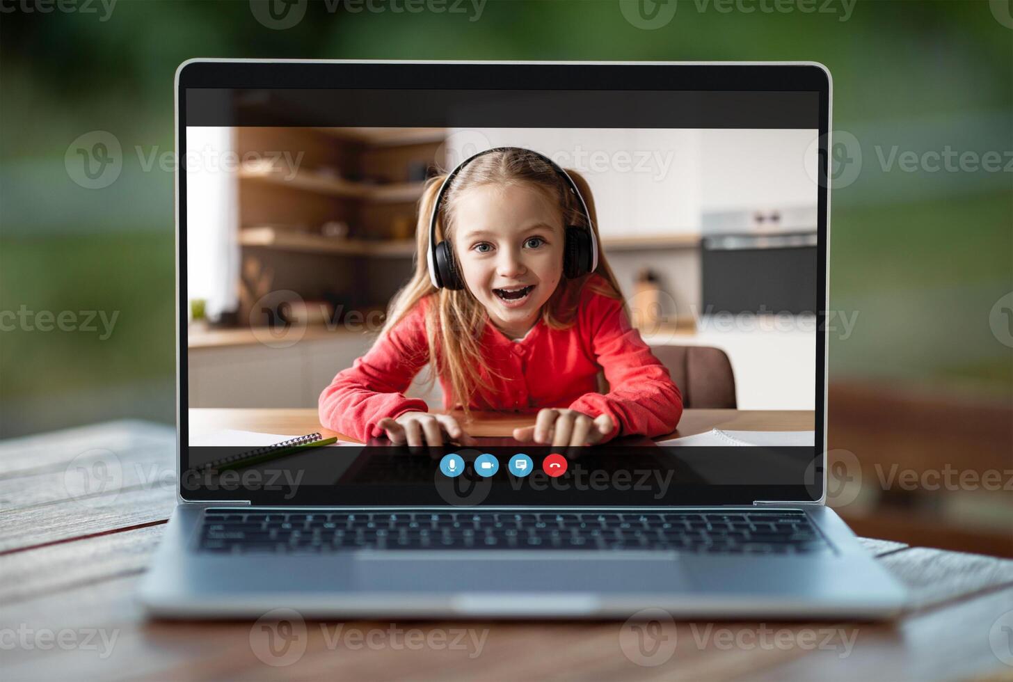 A young girl in headphones sits at a desk in front of a laptop, smiling and looking at the camera. She is likely participating in an online class or chat photo