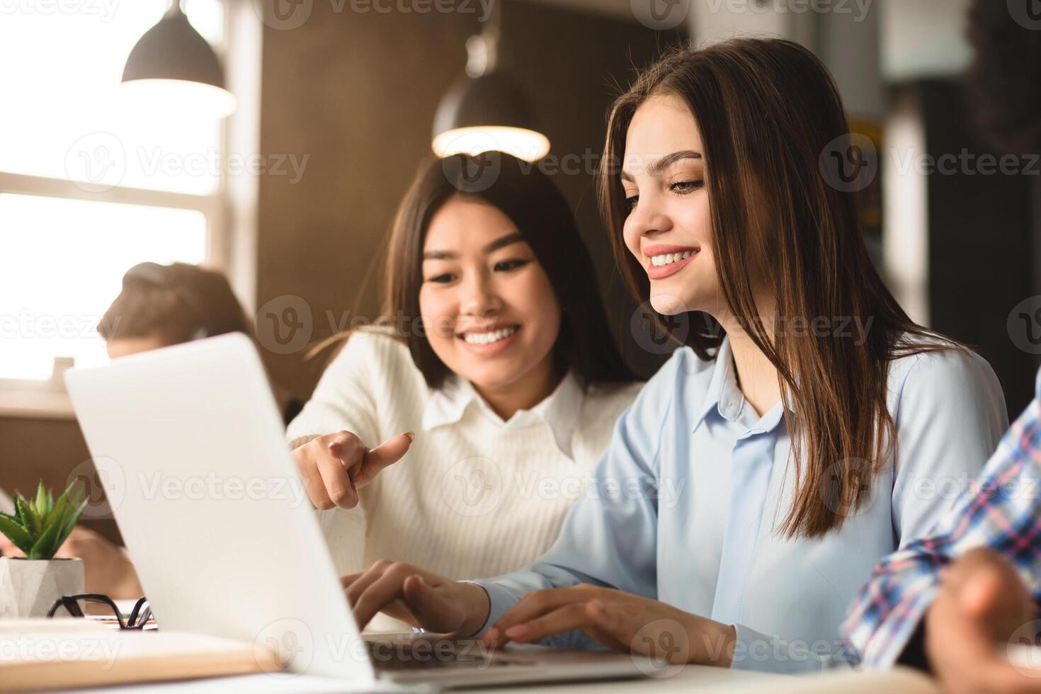 Two multiethnic women are seated at a desk in a well-lit library, working together on a project. One is pointing at a laptop screen, suggesting an idea or solution photo