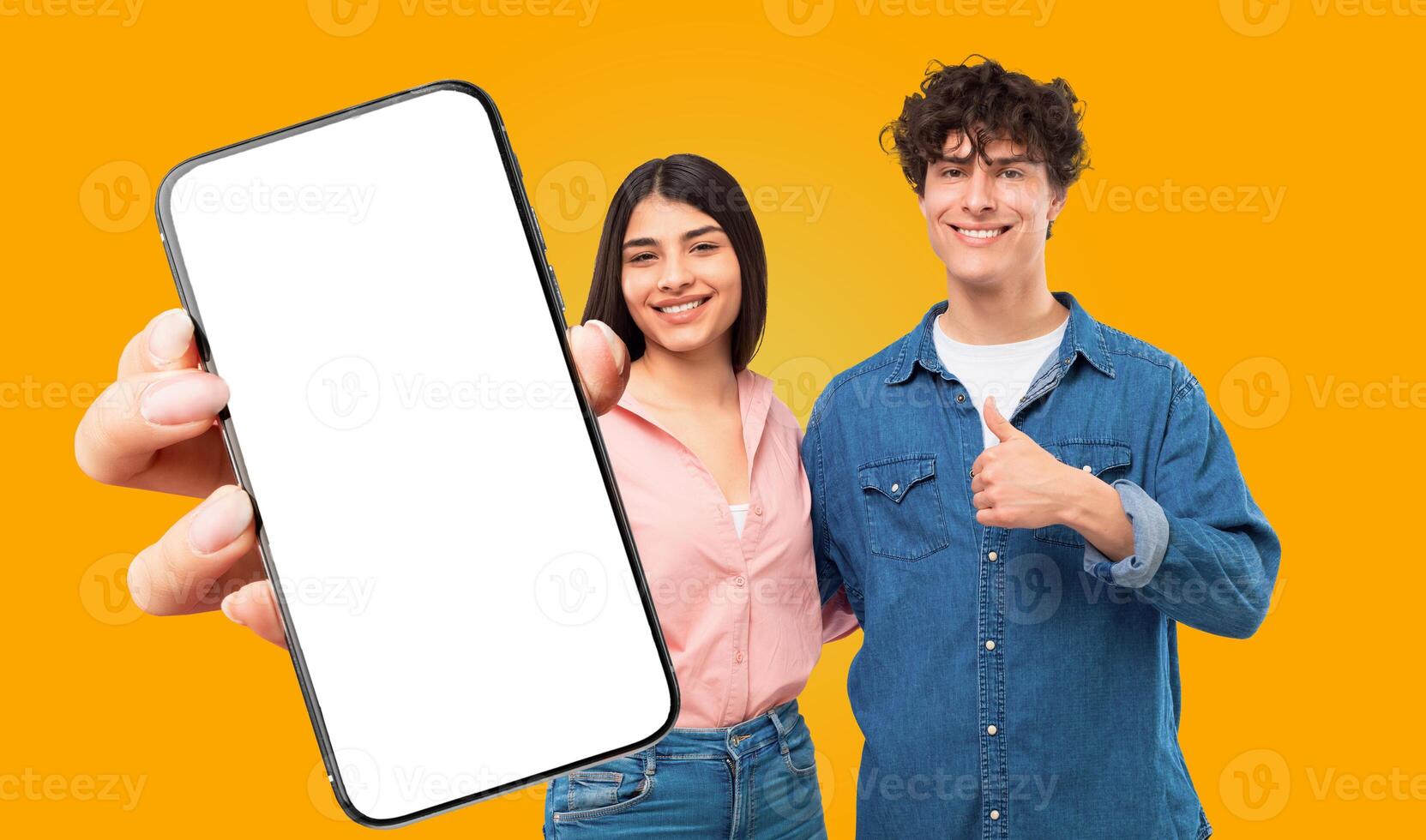 A young man and woman stand in front of a yellow background. The woman is holding a smartphone with a blank white screen towards the viewer, while the man gives a thumbs up. Both are smiling. photo