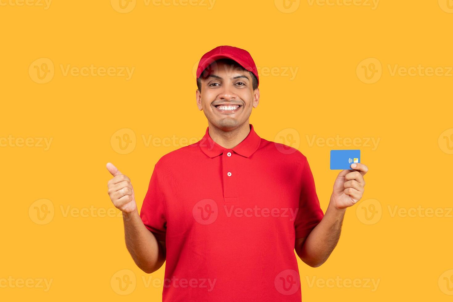 A cheerful delivery man dressed in a red uniform is giving a thumbs up gesture while holding a blue card. The background is a vibrant yellow, adding to the positive and energetic atmosphere photo