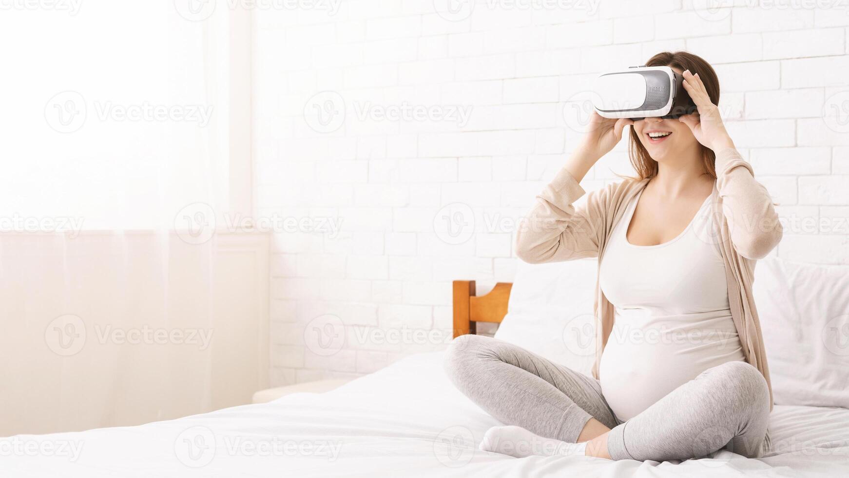 A pregnant woman is sitting on a bed, engrossed in using a virtual reality device. She appears focused and engaged with the technology, experiencing a virtual environment, copy space photo