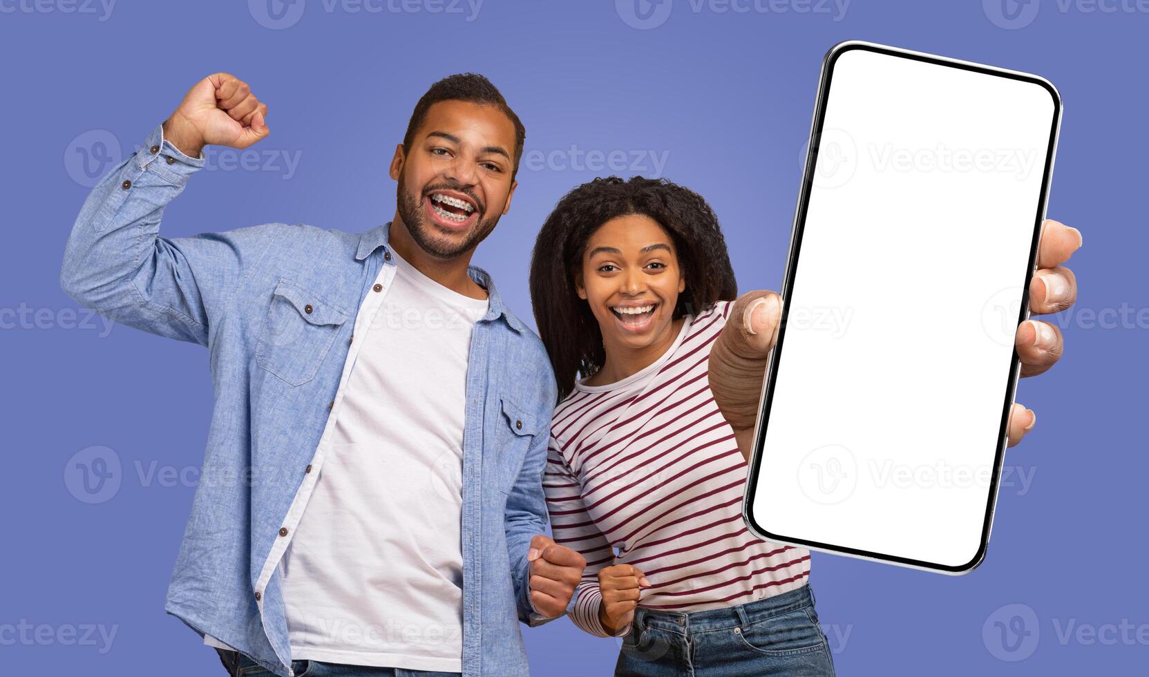 A young black couple stand against a purple backdrop. The woman holds a smartphone with a blank screen toward the camera, her finger pointing at it. The man stands with his arm raised photo