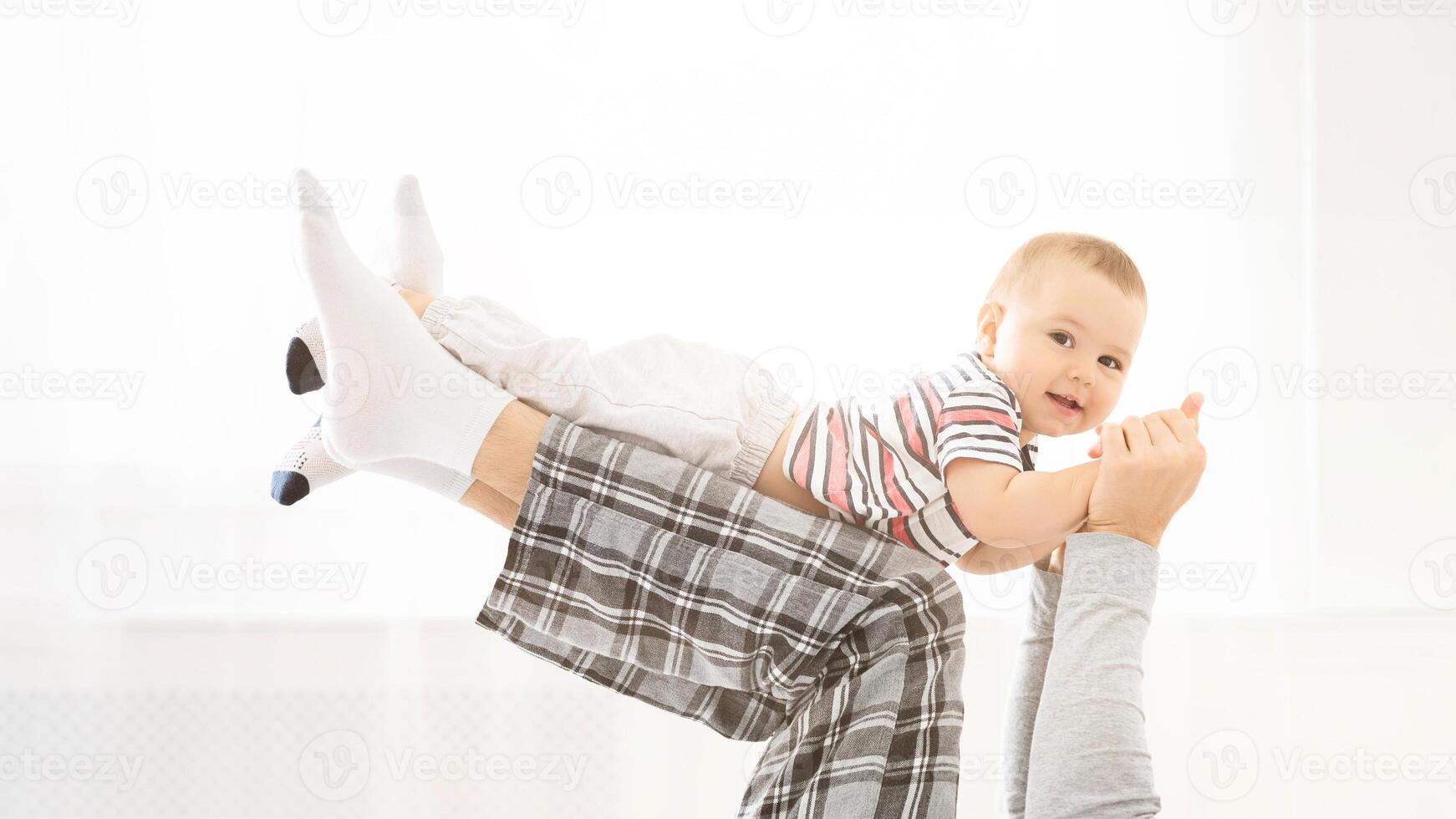 An image of a father holding his baby son in the air. The father is wearing plaid pajama pants and the baby is wearing a striped shirt. Both are smiling. photo