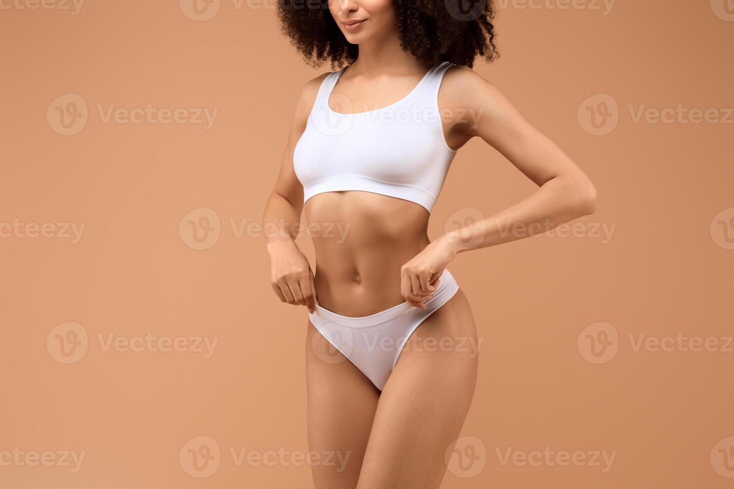 African American woman with brown hair stands against a peach-colored background. She is wearing a white sports bra and white underwear. She is looking at the camera, cropped photo