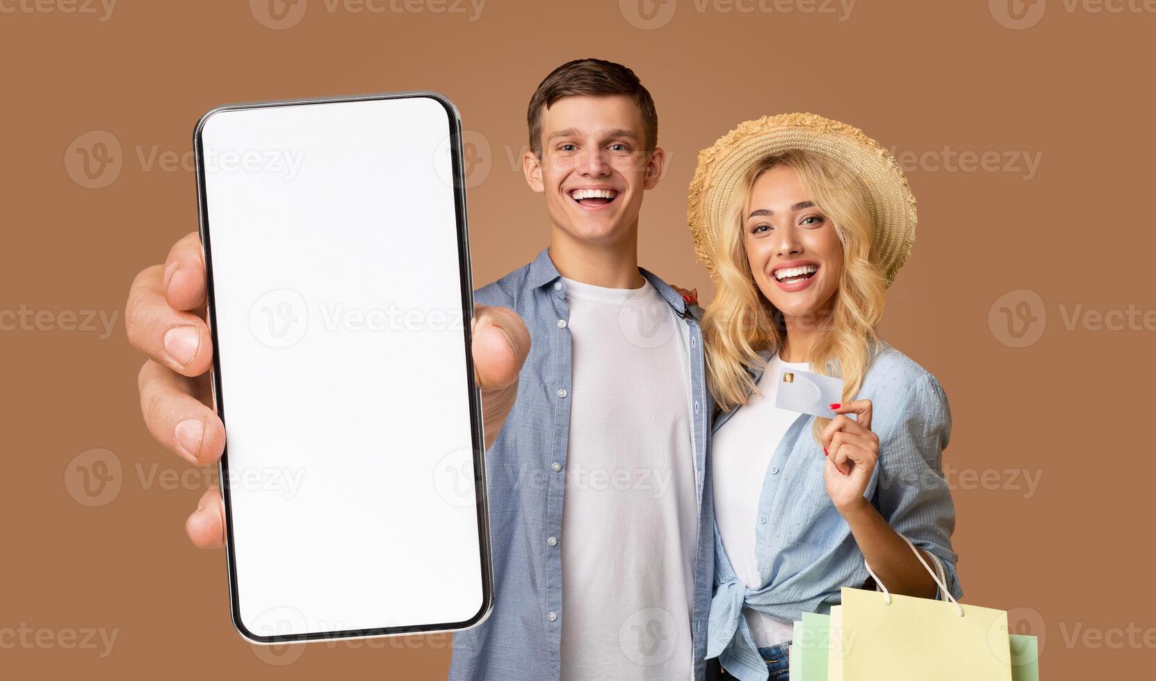 A young couple stand smiling in front of a brown background. The man is wearing a blue plaid shirt and is holding a smartphone with a blank white screen toward the camera photo