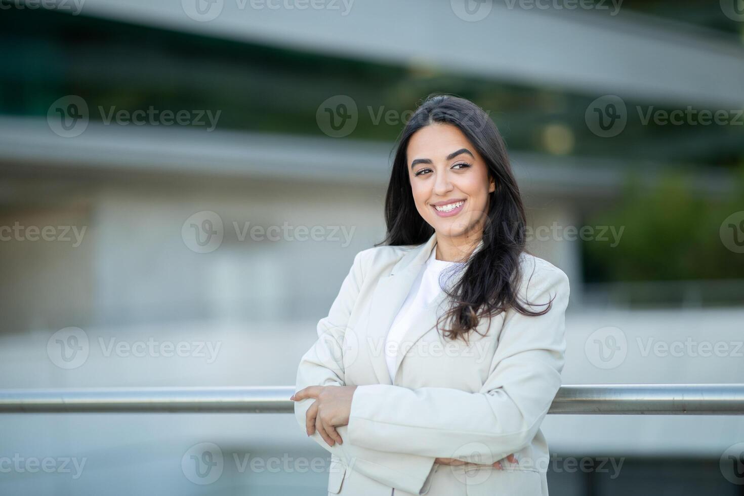 A young woman stands confidently with her arms crossed, smiling warmly, in an urban area with modern architecture. photo