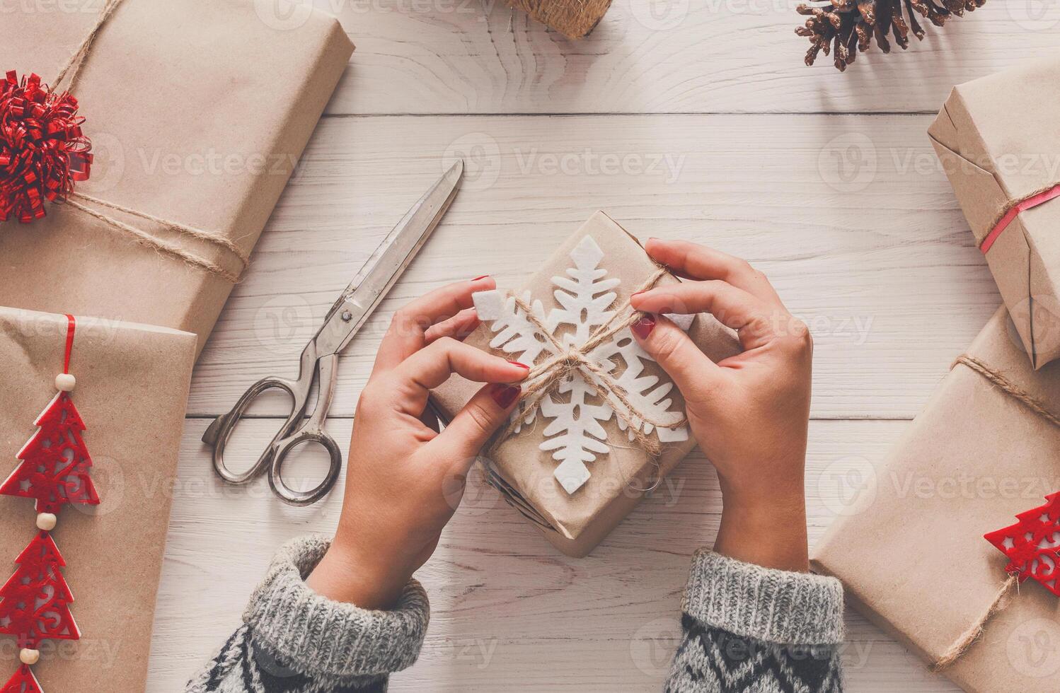 Creative hobby. Woman's hands wrap christmas holiday handmade present in craft paper with twine ribbon. Making bow at xmas gift box, decorated with snowflake. Scissors on white wooden table, top view. photo
