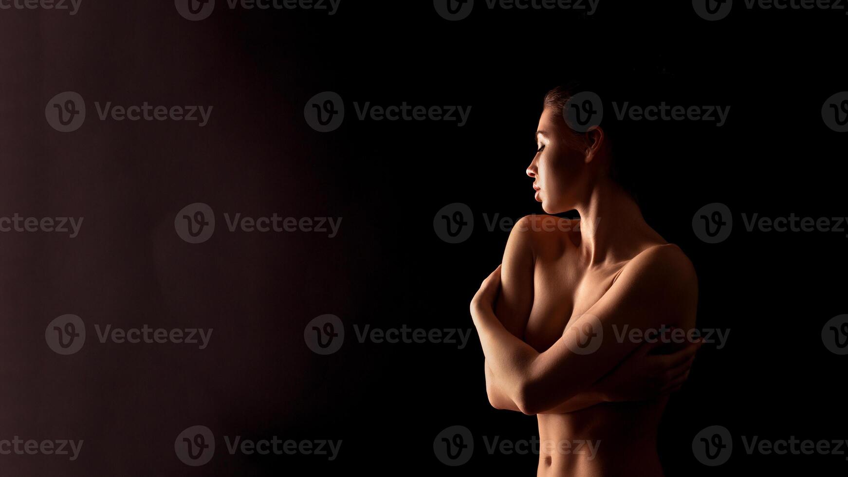 This image shows a woman standing in a dark room with her arms crossed over her chest. The lighting is soft and highlights her body and face in silhouette. photo