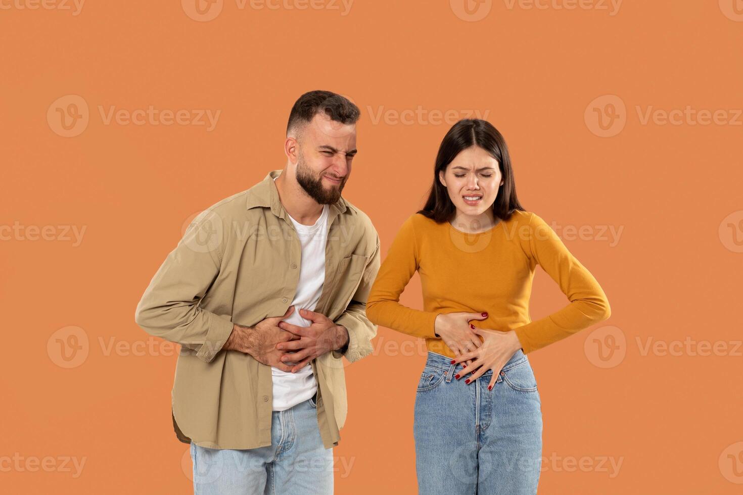 A young couple holds their stomachs, appearing to endure discomfort against an orange backdrop. Both are casually dressed, emphasizing their distress. photo
