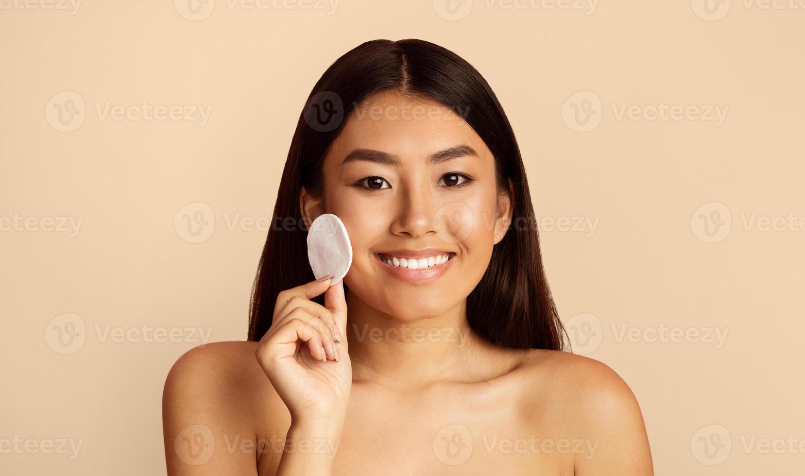 A young Asian woman with long black hair smiles at the camera while applying a cotton pad to her cheek as part of her skincare routine. The background is a simple, light beige color. photo