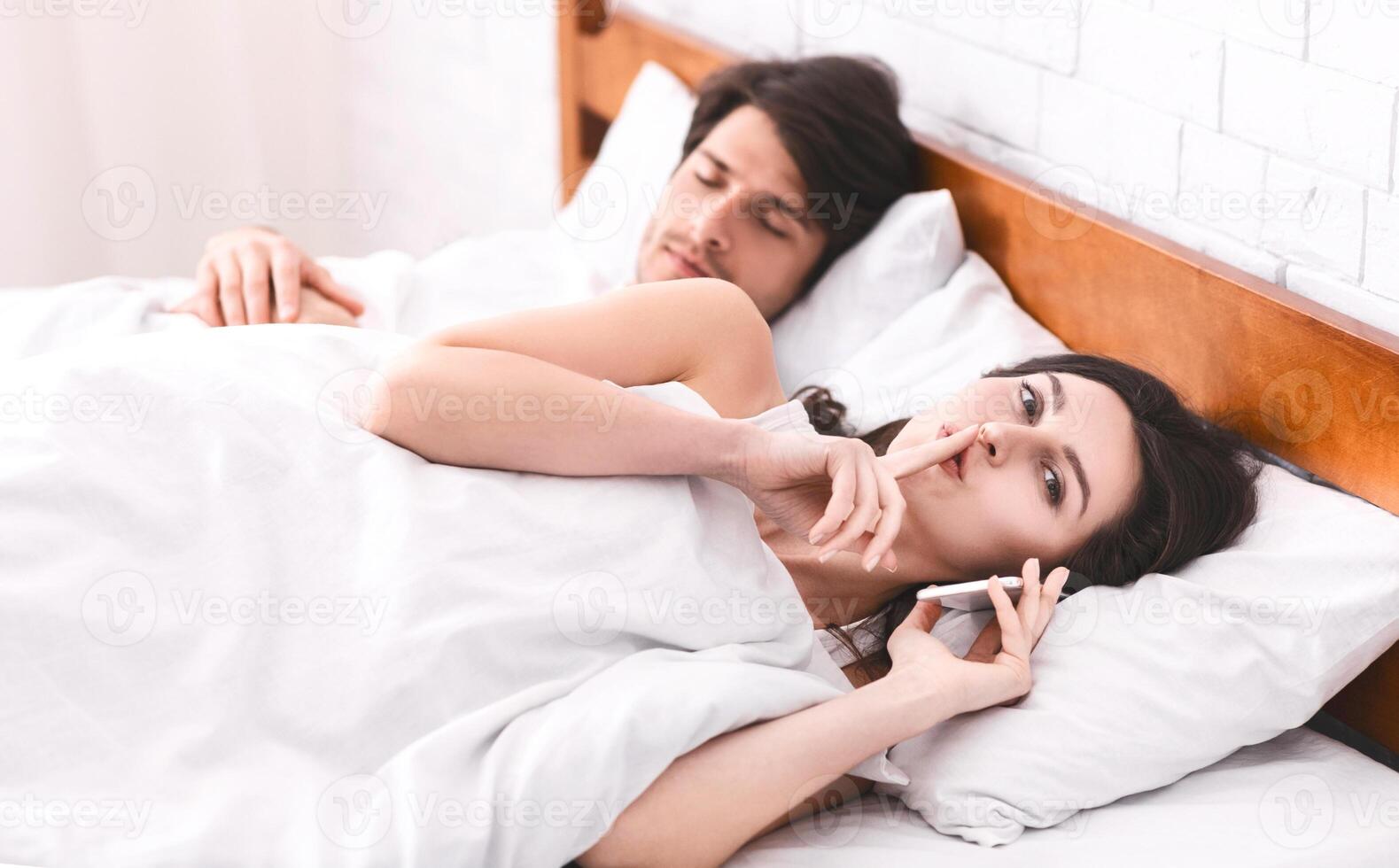 A woman is lying in bed next to a sleeping man, holding a phone to her ear while whispering into it with her finger on her lips. The man appears to be asleep, unaware of secretive phone call. photo