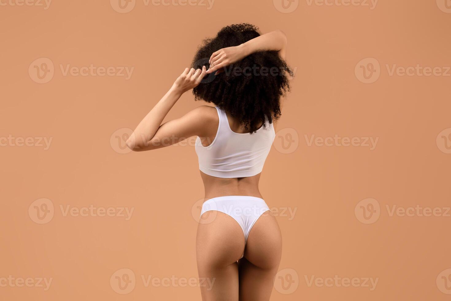 A woman with dark curly hair is standing facing away from the camera in a light brown room, wearing a white tank top and white underwear, with her right arm raised behind her, touching her hair. photo