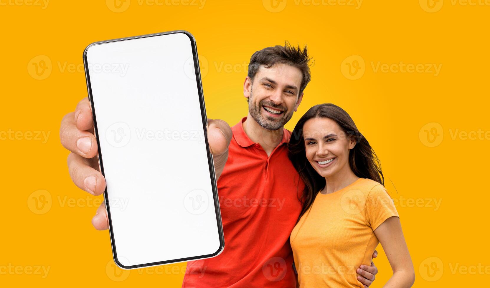 A young couple smiles at the camera while holding a smartphone with a blank screen in front of a bright yellow background. photo