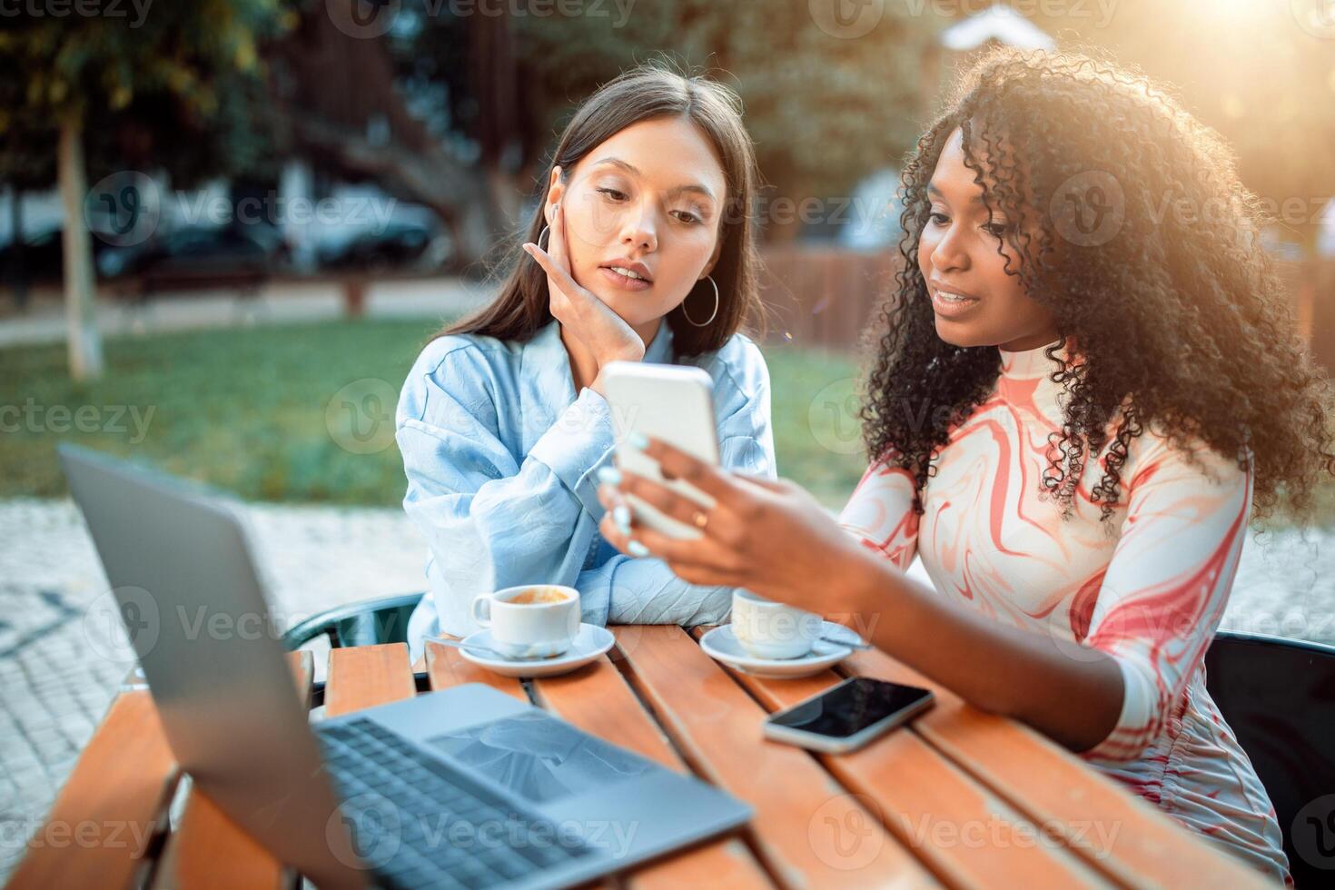 This image shows two women seated at a table outside of a cafe. One of the women is showing something on her smartphone to the other. They using laptop, working on project photo