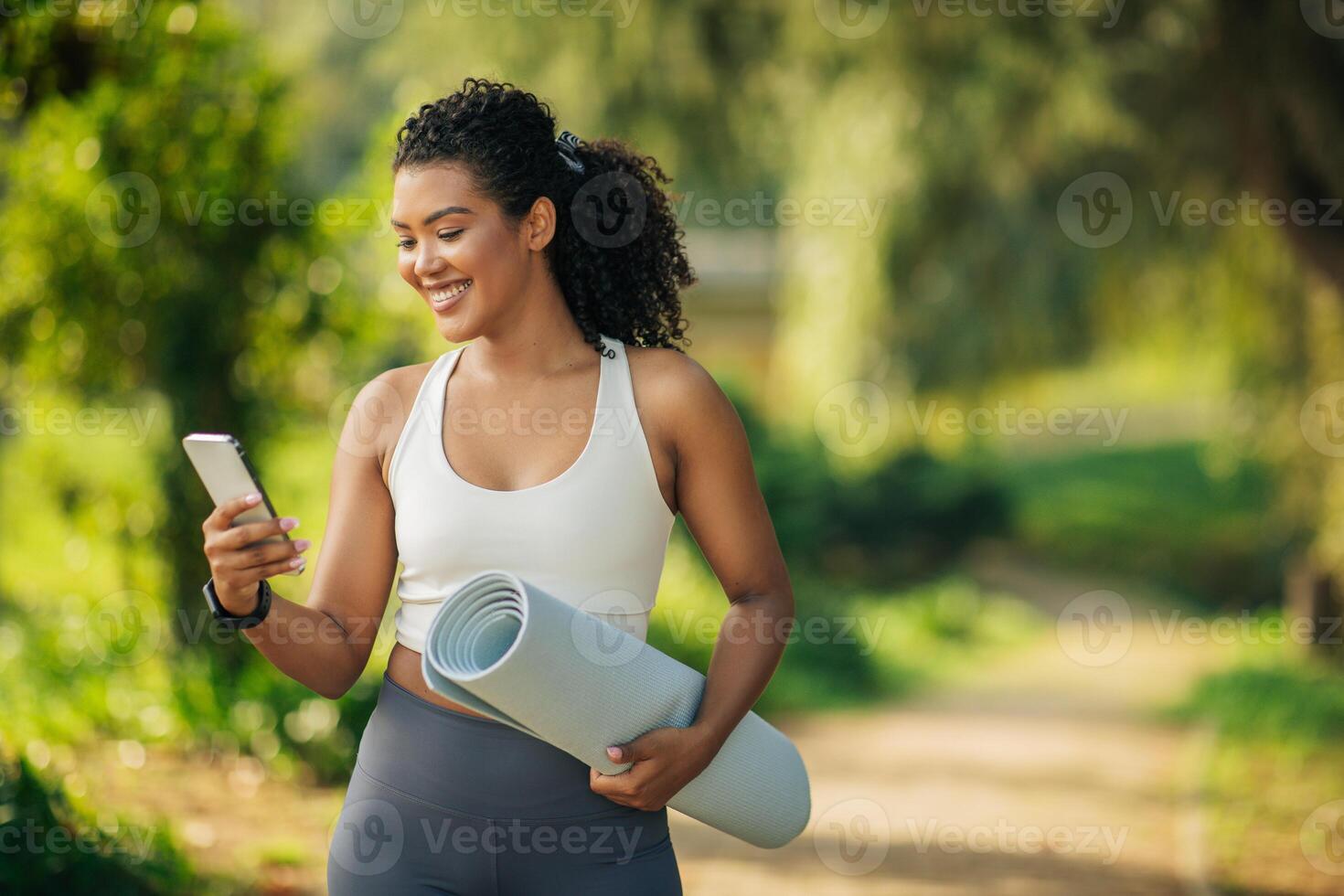 A smiling woman with curly hair is walking in a park while holding a rolled-up yoga mat in her right hand and using her smartphone with her left hand, copy space photo