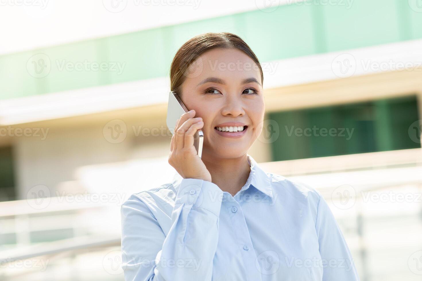 A young Asian woman with a cheerful expression is engaged in a phone conversation while standing outdoors. She is dressed in a light blue shirt and appears to be enjoying her discussion. photo