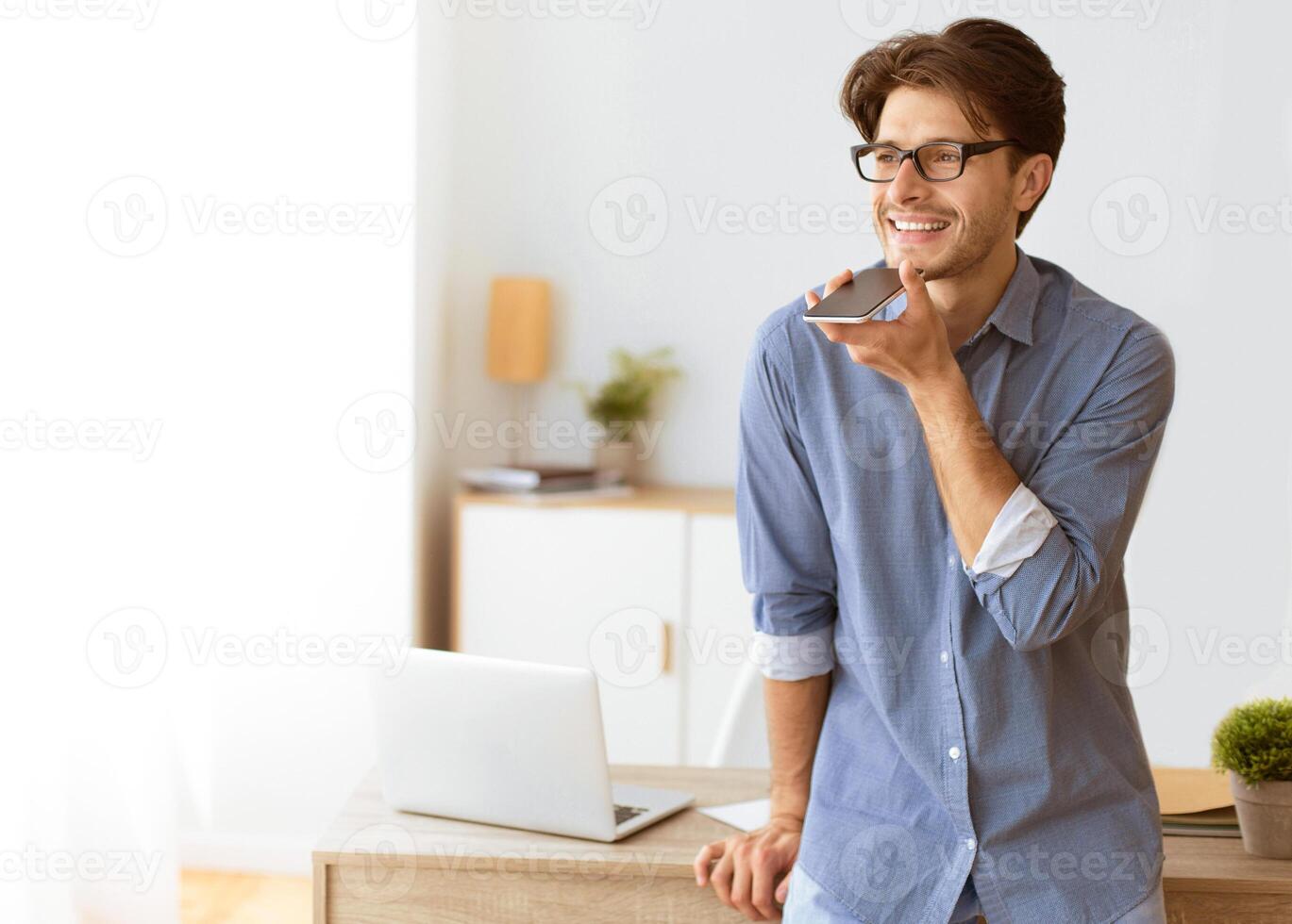 A man wearing glasses stands in his office, smiling while using a voice assistant on his phone. He is leaning against a desk with a laptop and other office supplies. photo