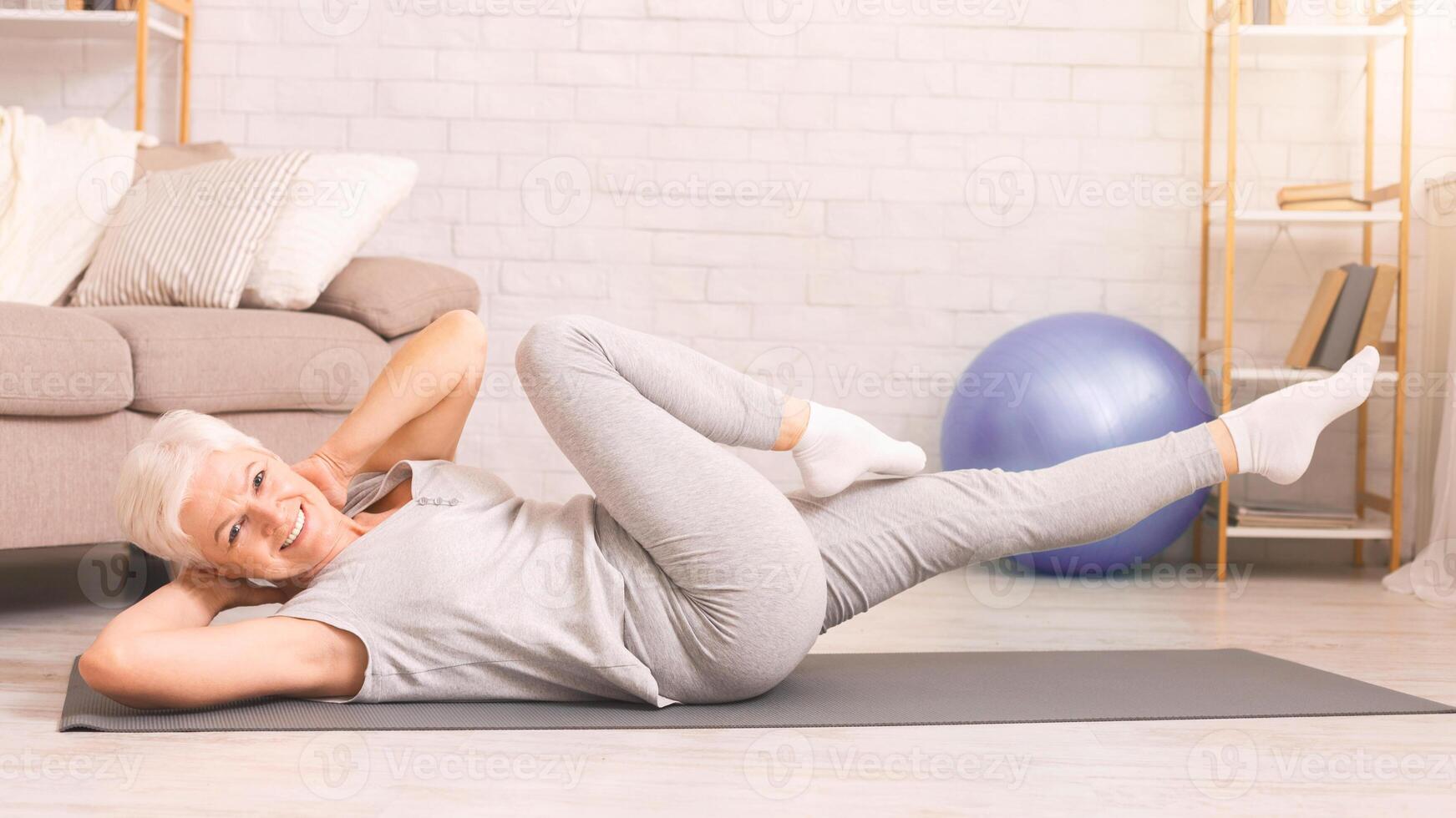 Senior woman is engaged in a yoga pose on a mat placed on the floor. She appears focused and concentrated, displaying flexibility and strength in her body movements. photo