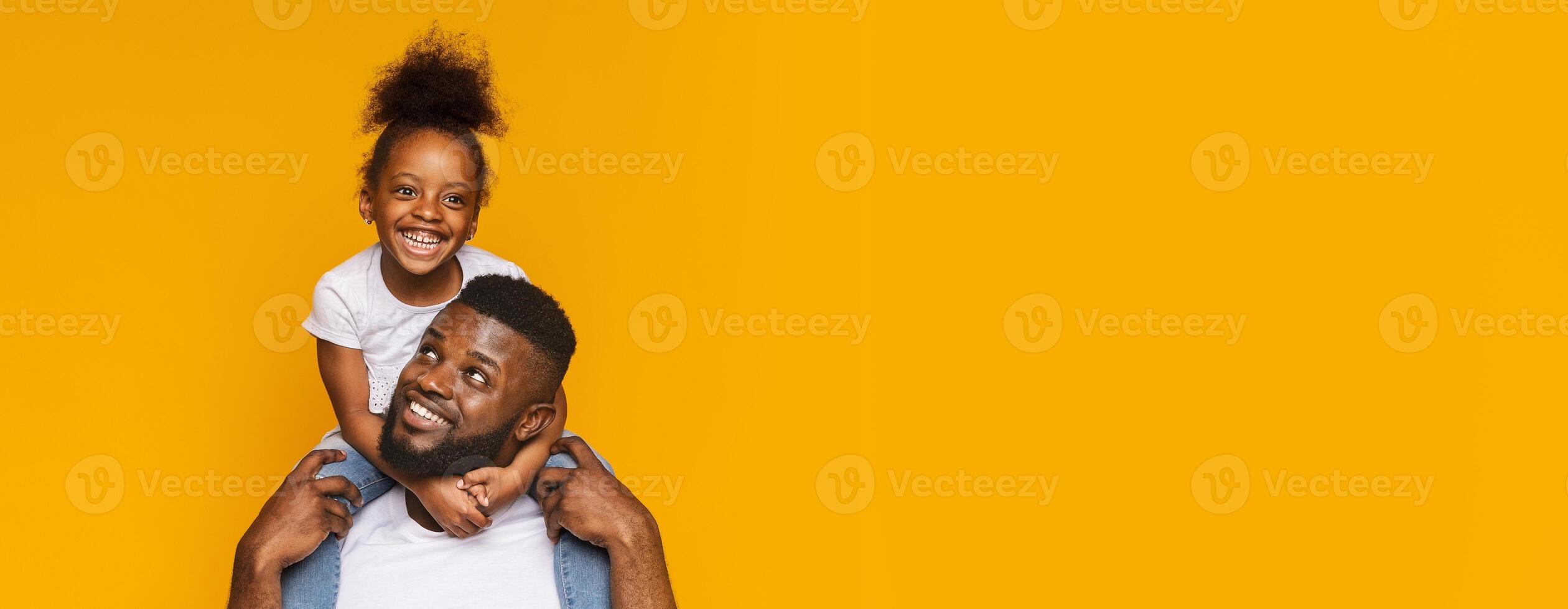 African American father happily carries his daughter on his shoulders, both beaming with joy. They are set against a vibrant yellow backdrop that enhances their cheerful expressions, copy space photo