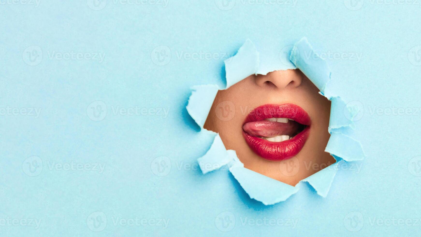 A woman mouth is visible, peeking out of a hole in a piece of paper. The woman lips with red lipstick are slightly parted, creating a mysterious and intriguing sight, copy space photo