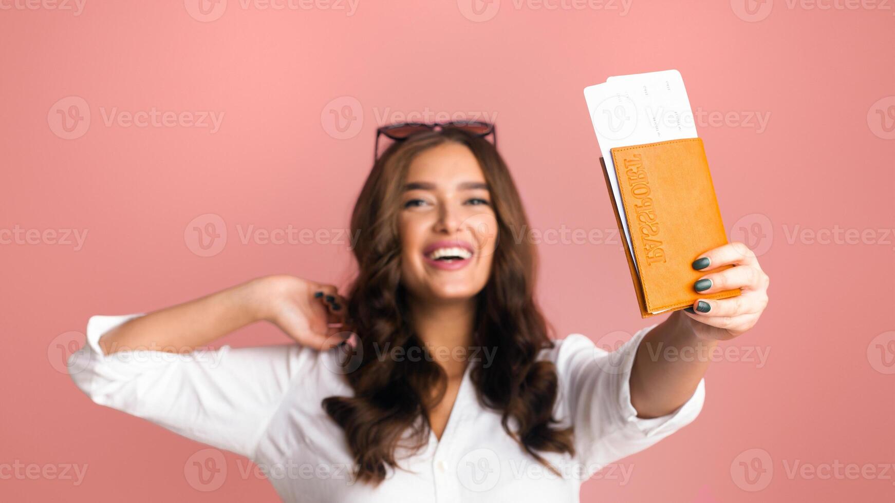 A young woman with wavy hair and sunglasses pushed up on her head is joyfully showing passport with flight tickets. She is seen in a carefree pose, wearing a white shirt, against a soft pink backdrop photo