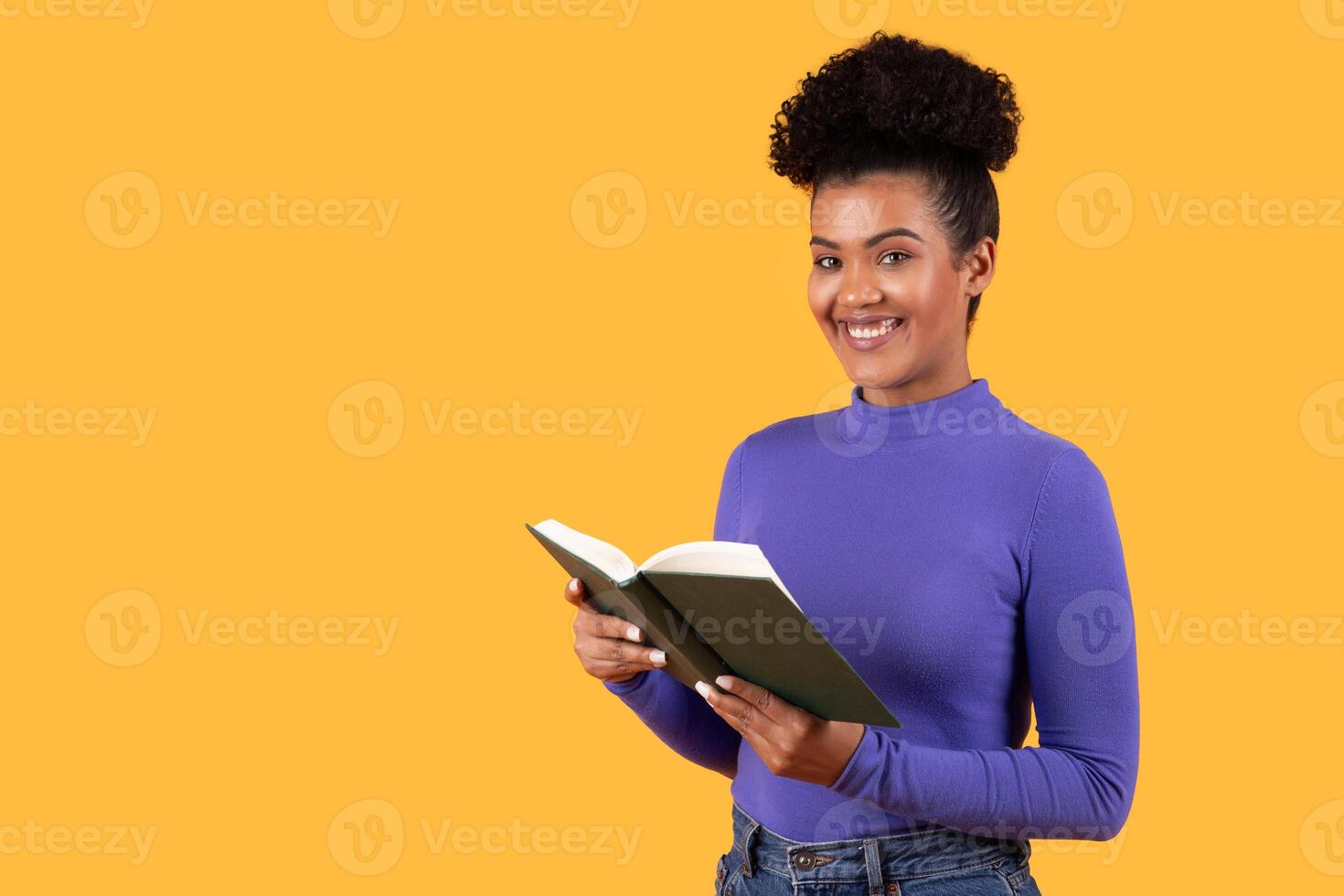 This image shows a young woman with dark skin and curly hair, smiling while holding a book open in front of her. She is wearing a purple shirt and jeans and is standing in front of yellow background. photo
