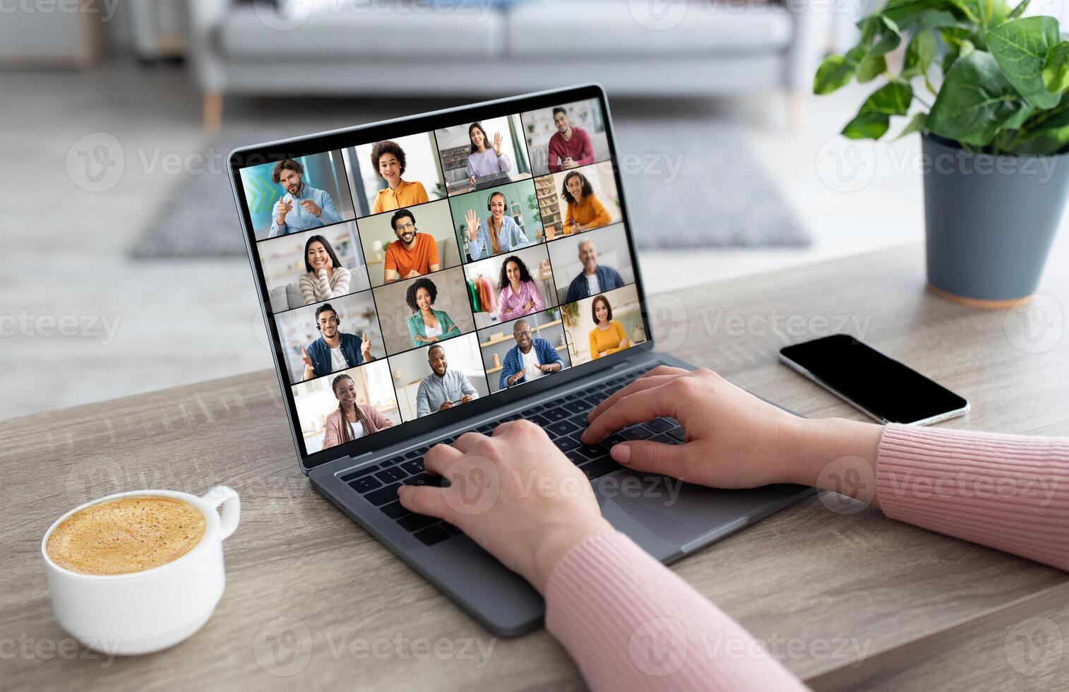 Woman sits at a wooden table with a laptop in front of her. The laptop screen displays a conference with many participants. The lady is typing on the laptop keyboard with one hand photo