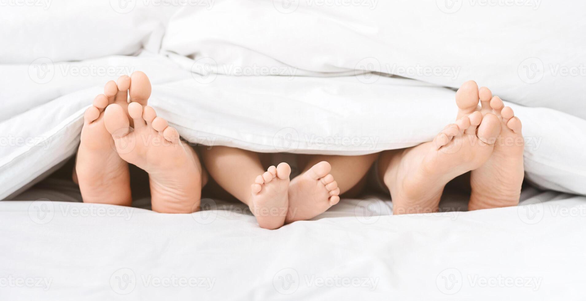 A close-up photo of a couple and baby feet sticking out from under the covers of a white bed. The feet are bare and relaxed, suggesting a comfortable and intimate setting.