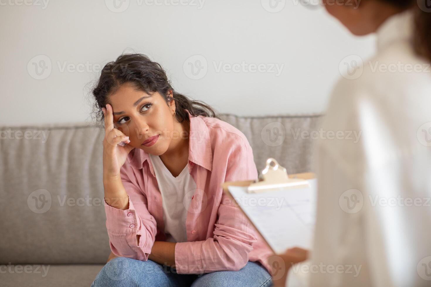 A woman sits on a couch during a therapy session. She has her chin on her hand and looks down, appearing thoughtful or troubled. The therapist stands out of view, holding a clipboard with paperwork. photo