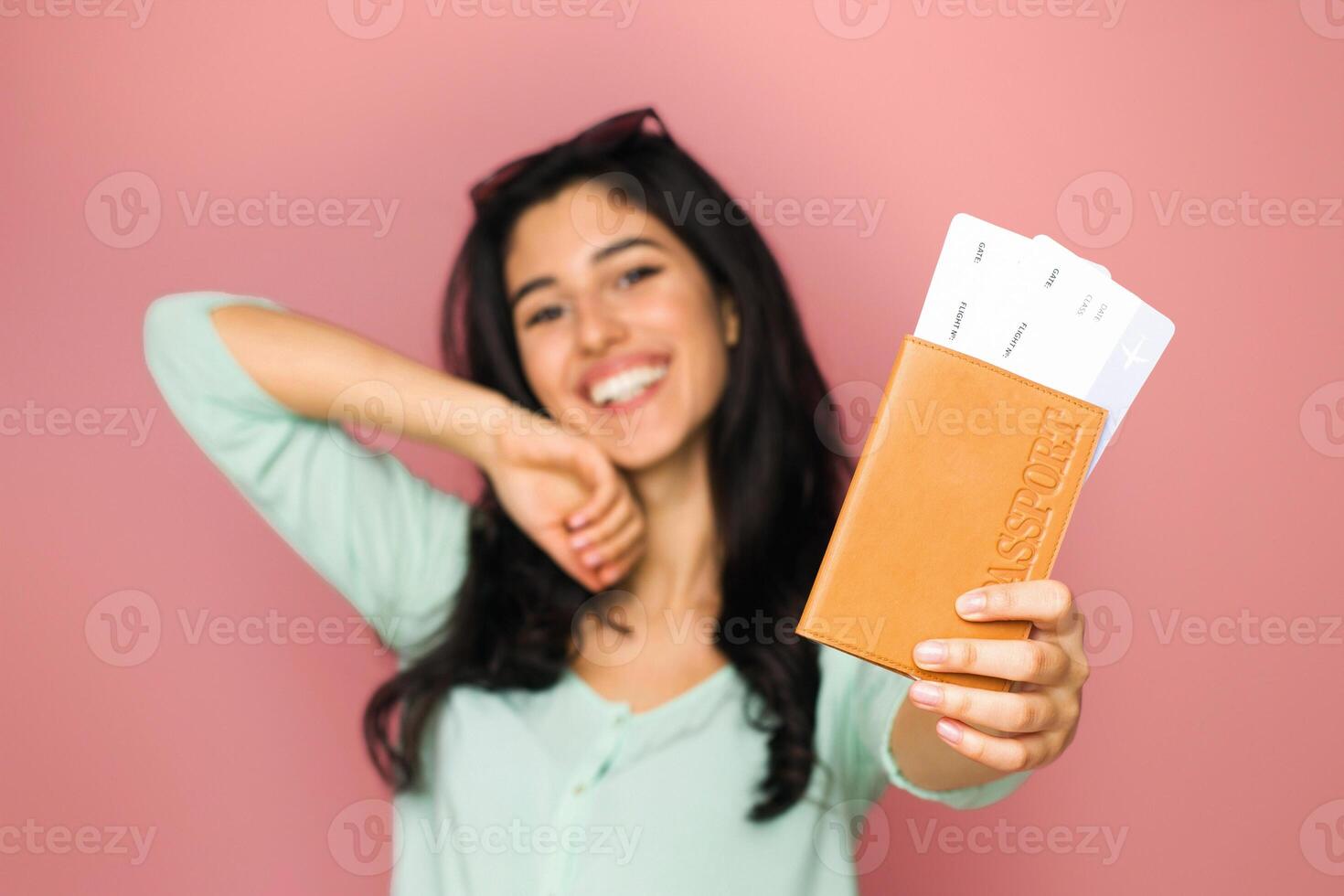 A cheerful young middle eastern woman stands against a soft pink backdrop, proudly showing off her passport and boarding pass. photo