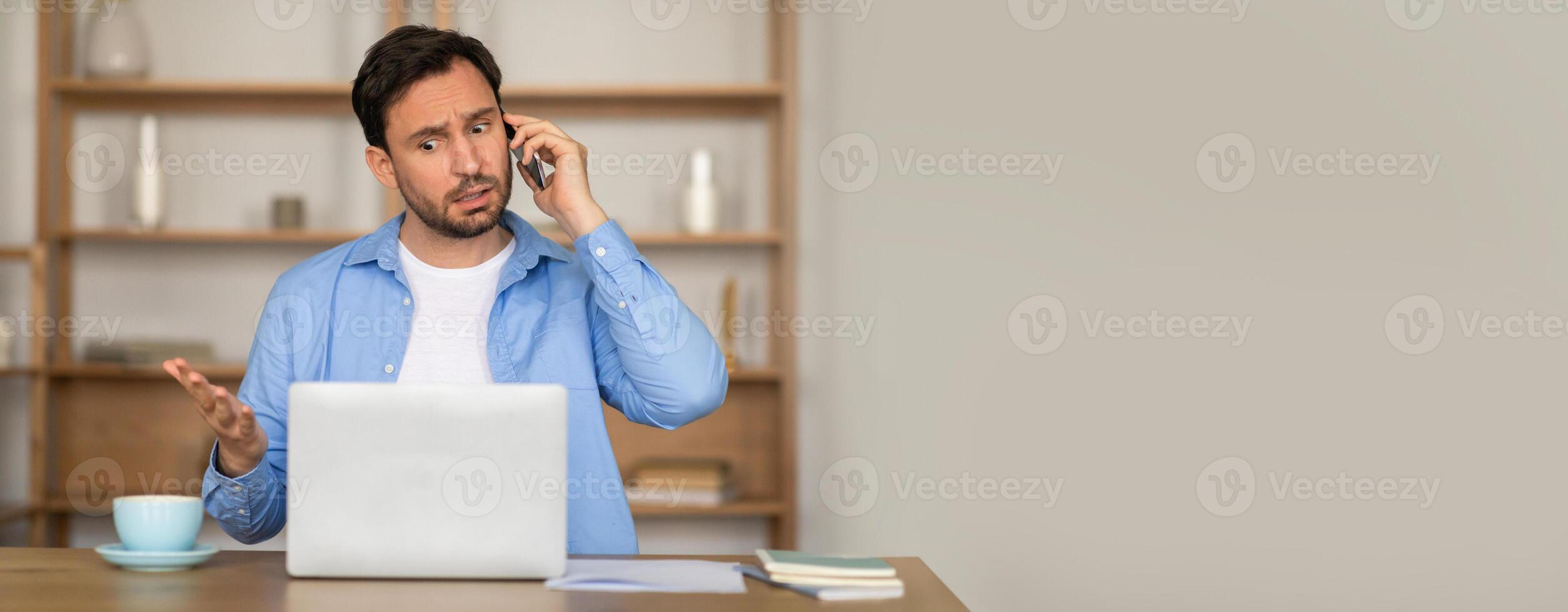 A man dressed casually engages in a phone conversation while sitting at his desk with a laptop open. He appears focused and slightly frustrated, taking notes beside a coffee cup. photo