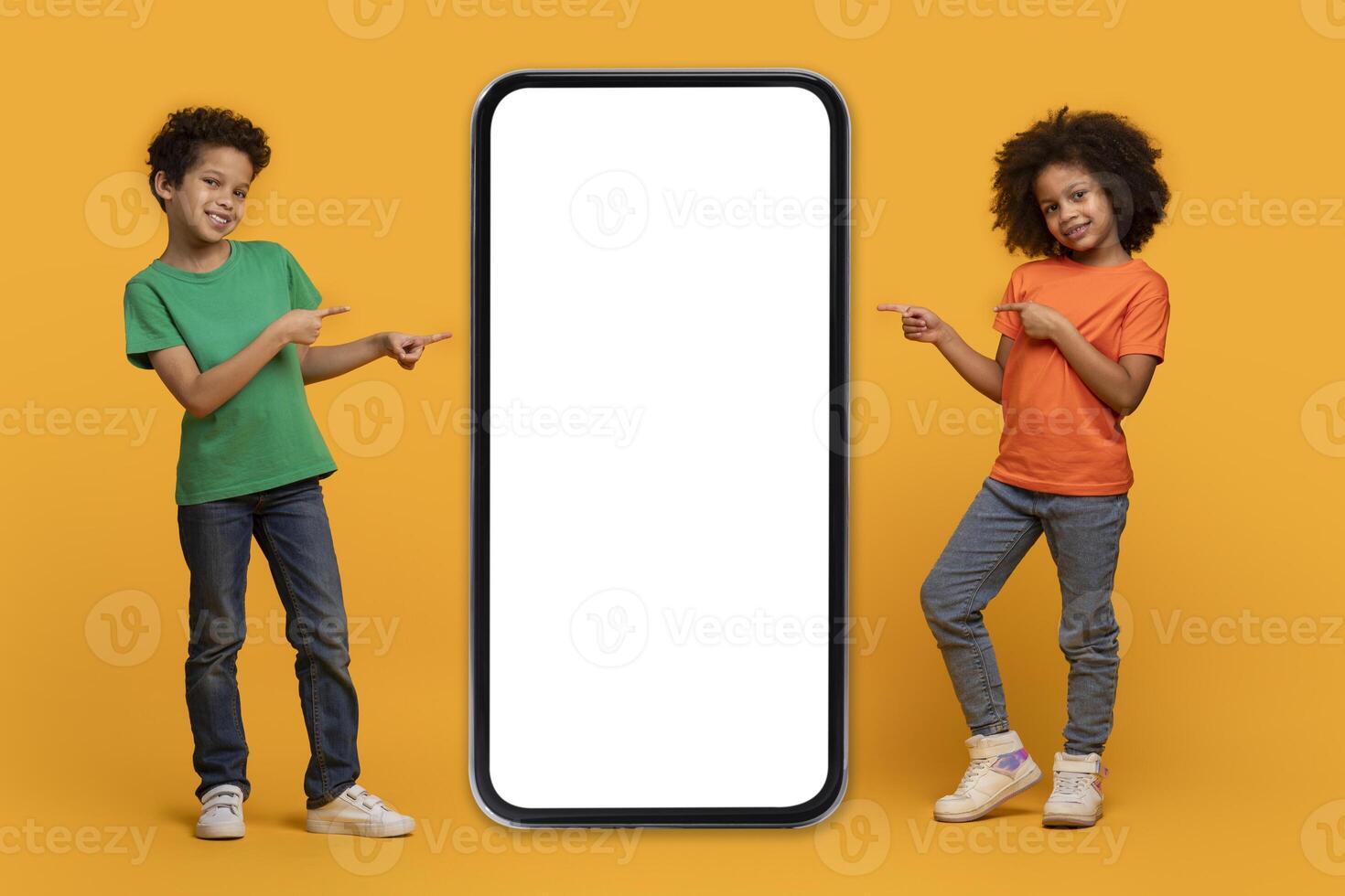 Two happy African American kids are standing on an orange backdrop, each pointing at a large blank screen of a vertically positioned smartphone, conveying a sense of excitement or presentation photo