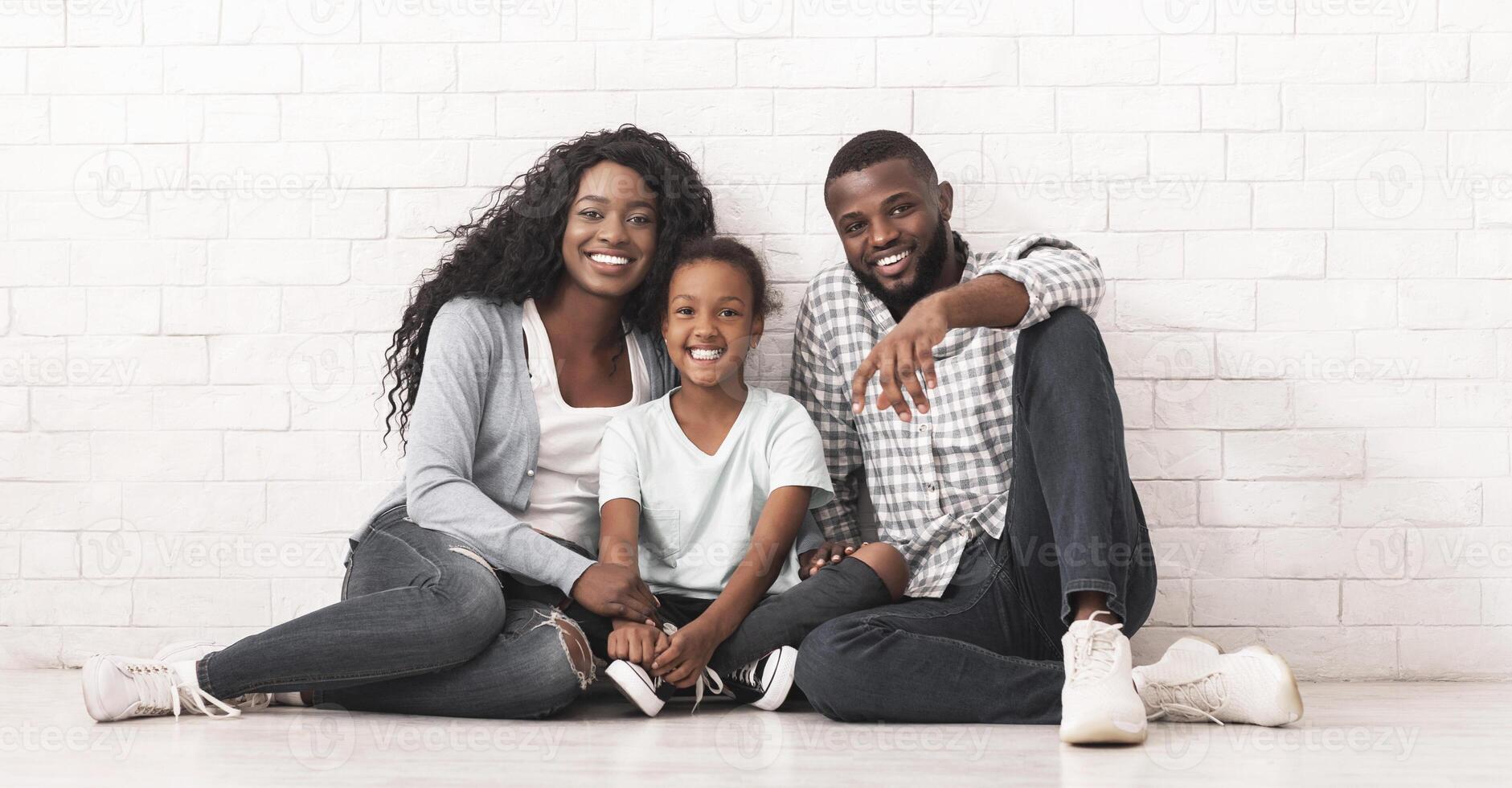 Happy Millennial Family Concept. Smiling Black Parents With Little Daughter Sitting On Floor Together, Smiling And Posing To Camera Over White Brick Wall Background, Free Space photo
