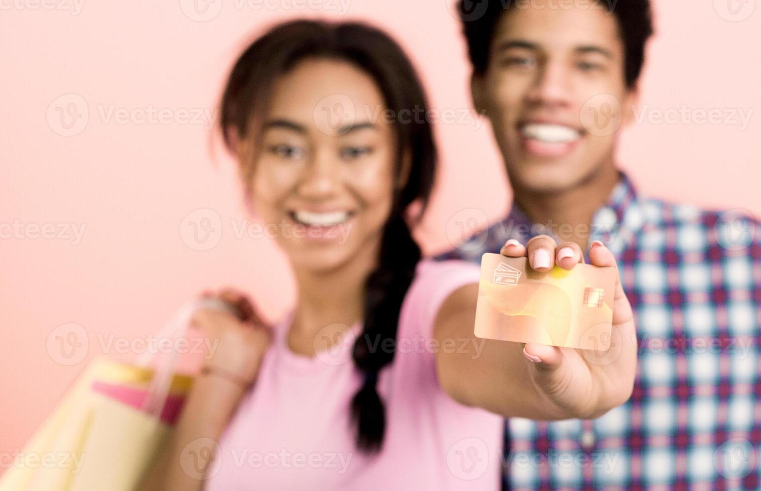 A young African American couple stands together, holding a credit card in front of a vibrant pink background. They both appear focused and engaged, possibly discussing financial decisions photo