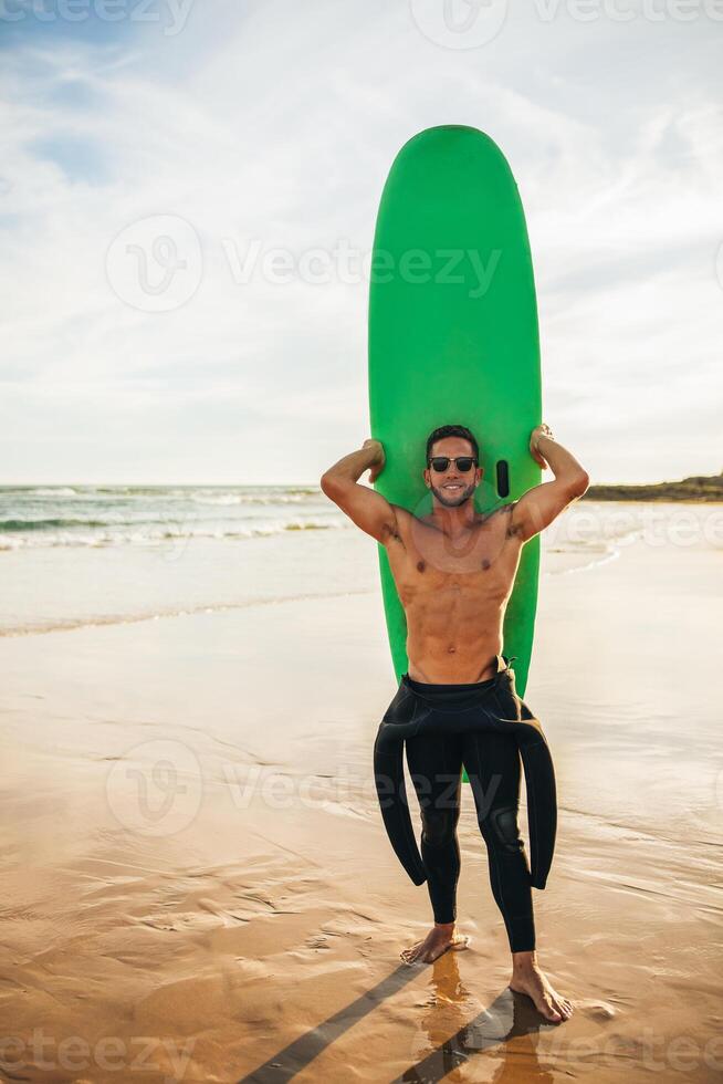 A man in a wetsuit is holding a green surfboard on a sandy beach. He is wearing sunglasses and smiling. The ocean is in the background and the sky is clear. photo