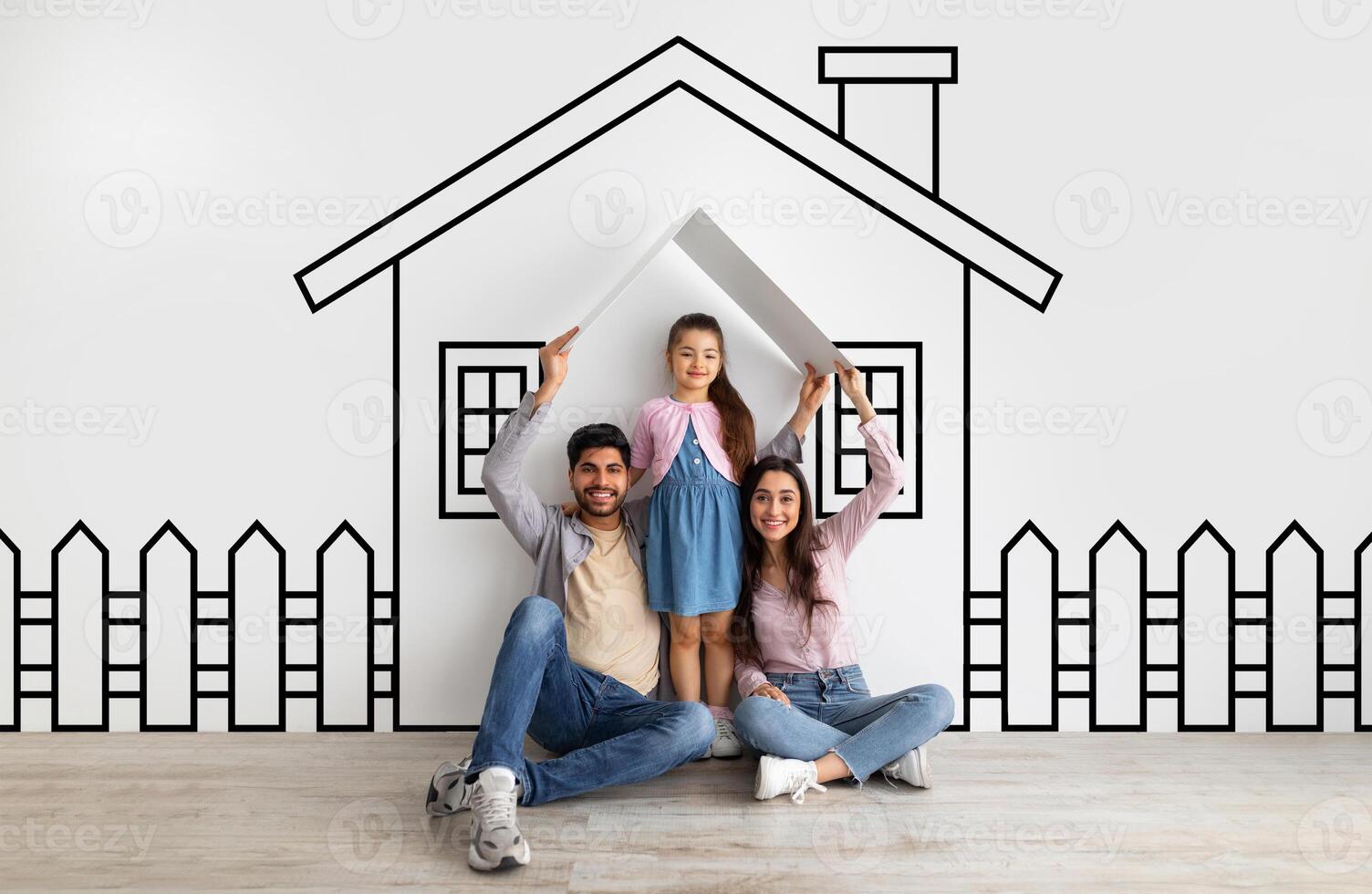 Happy middle eastern family father mother and daughter sitting on floor over illustrated house of their dreams over white wall background, parents holding a roof photo
