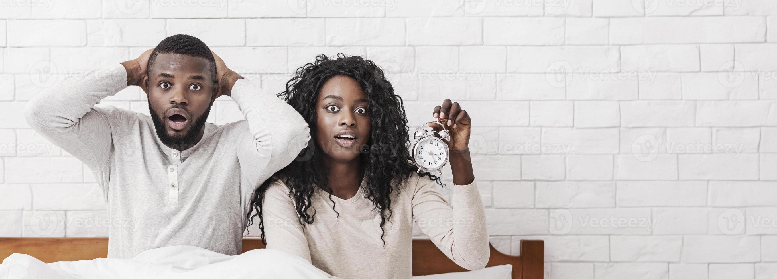 Oversleeping. Panicked black couple missed alarm clock ringing and sitting in bed with desperate face expression, free space photo